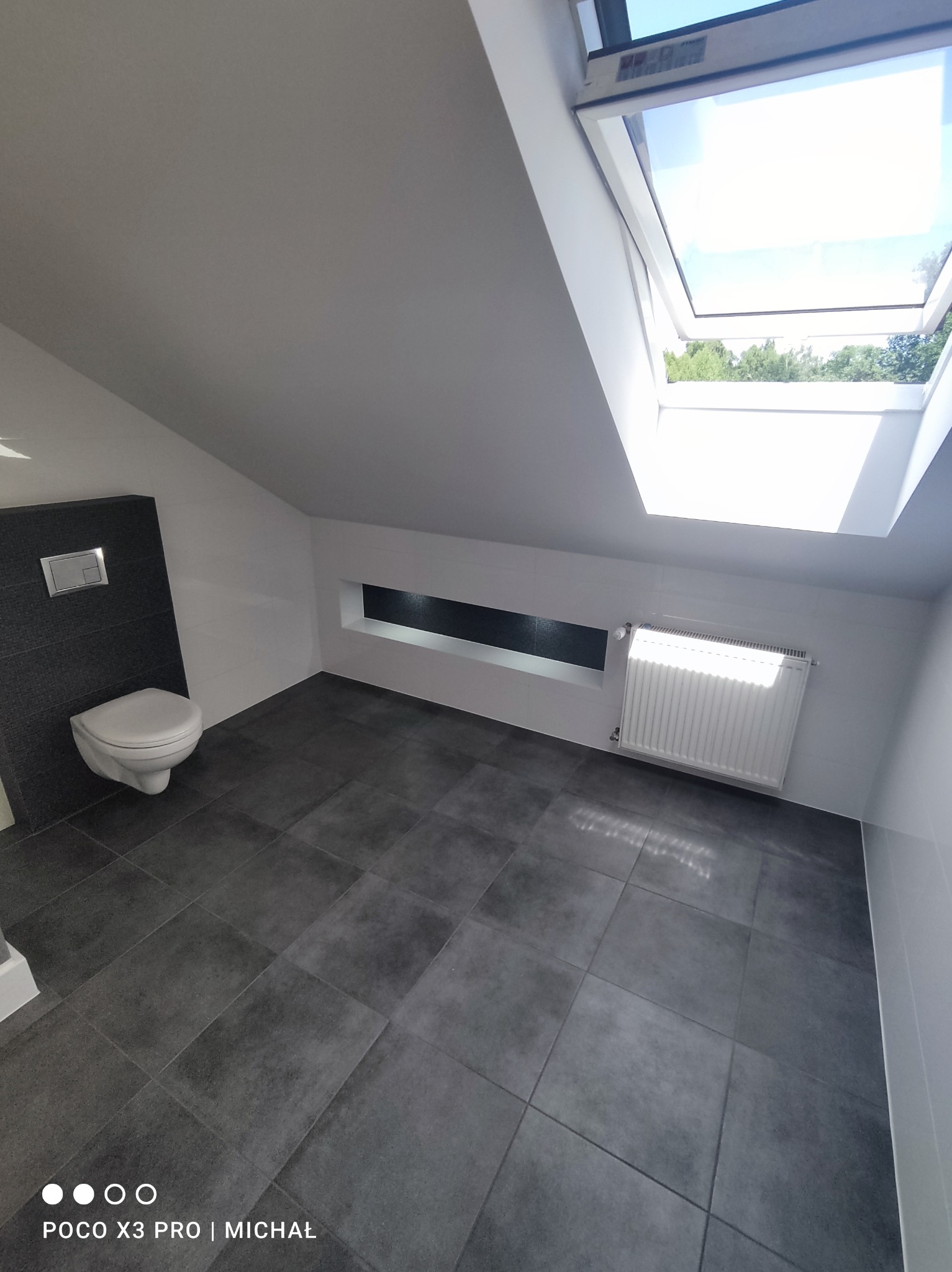 Nowoczesna łazienka na poddaszu z oknem dachowym, szarą glazurą podłogową i wiszącą toaletą na tle ciemnej ściany. Minimalistyczny design, białe ściany i grzejnik.