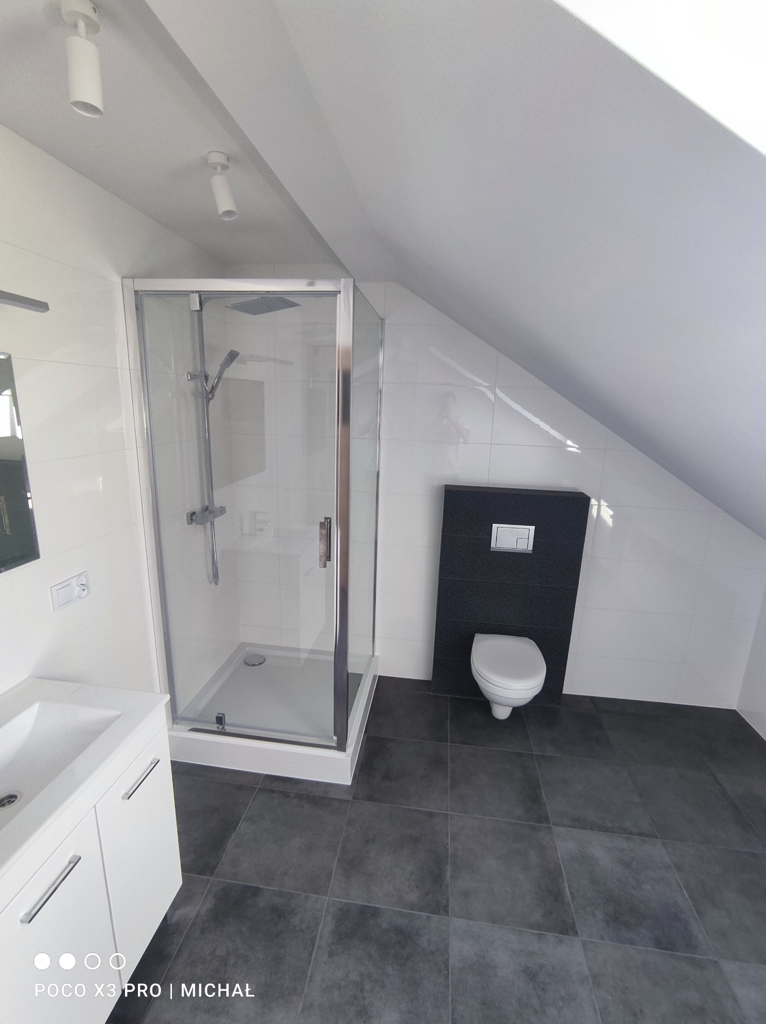 Nowoczesna łazienka na poddaszu z kabiną prysznicową, umywalką z szafką i toaletą. Białe płytki ścienne kontrastują z ciemną podłogą i zabudową toalety.