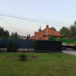 Świeżo wycięte drzewo w trawniku przed domem, widoczny pień po wycince, na podjeździe stoi biała ciężarówka z zielonym napisem 'Wycinka i pielęgnacja drzew SZYMPEK'.