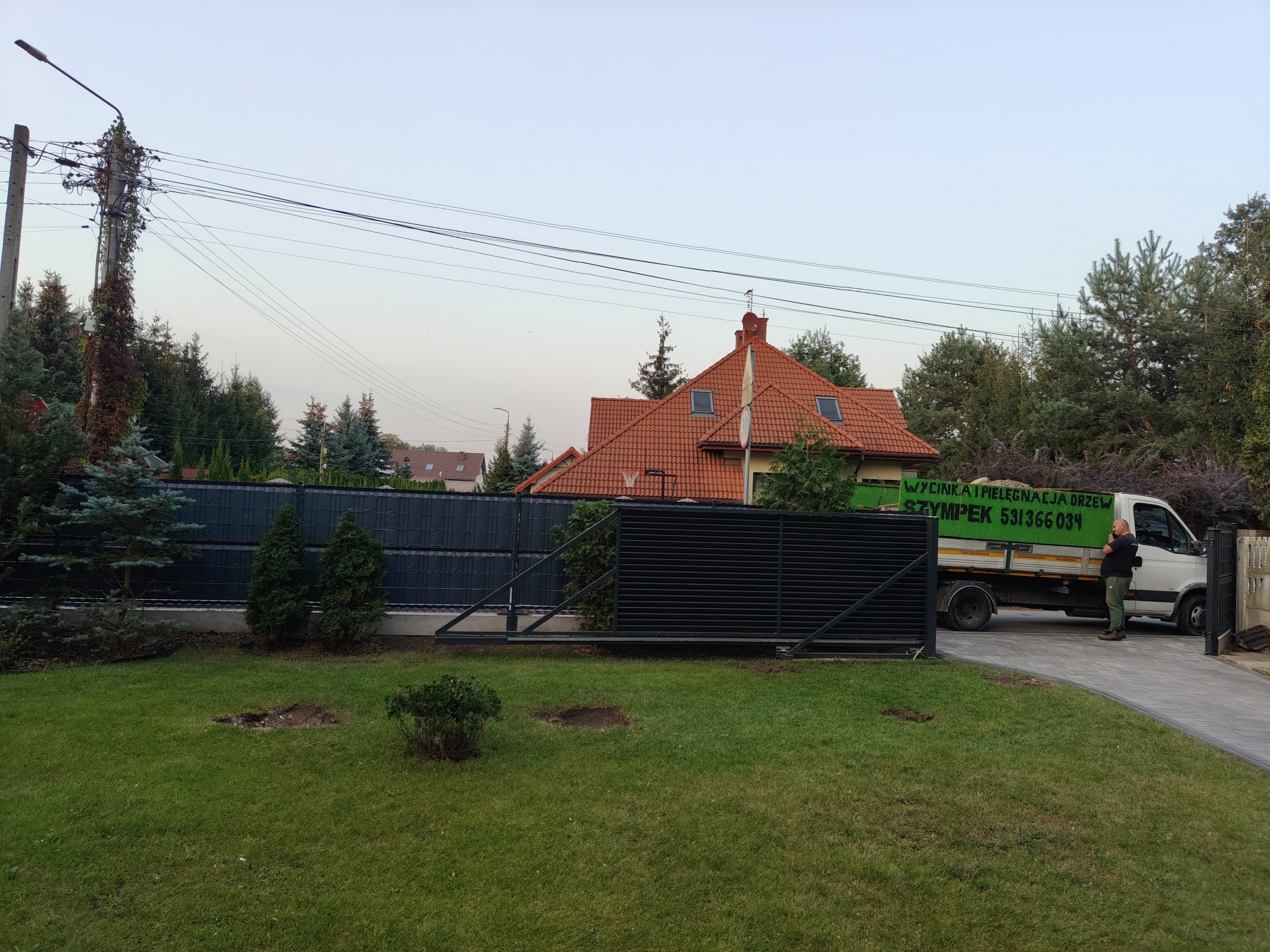 Świeżo wycięte drzewo w trawniku przed domem, widoczny pień po wycince, na podjeździe stoi biała ciężarówka z zielonym napisem 'Wycinka i pielęgnacja drzew SZYMPEK'.