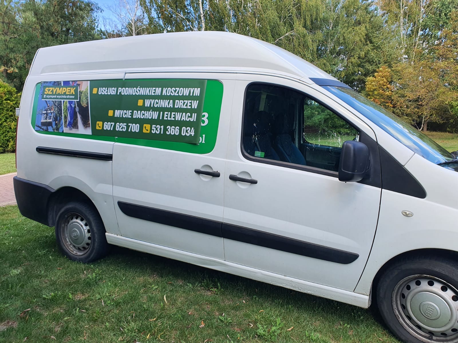 Biały samochód dostawczy firmy oferującej usługi z zakresu pielęgnacji zieleni, z reklamą na boku, zaparkowany na trawniku.