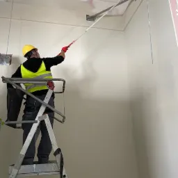 Pracownik w kasku i kamizelce odblaskowej maluje sufit hali przemysłowej za pomocą wałka na teleskopowym wysięgniku, stojąc na aluminiowej drabinie.