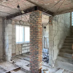 Wnętrze w trakcie remontu z widocznym słupem z cegły, stalowymi podciągami pod stropem i schodami, ukazujące etap prac po wyburzeniu ściany nośnej.