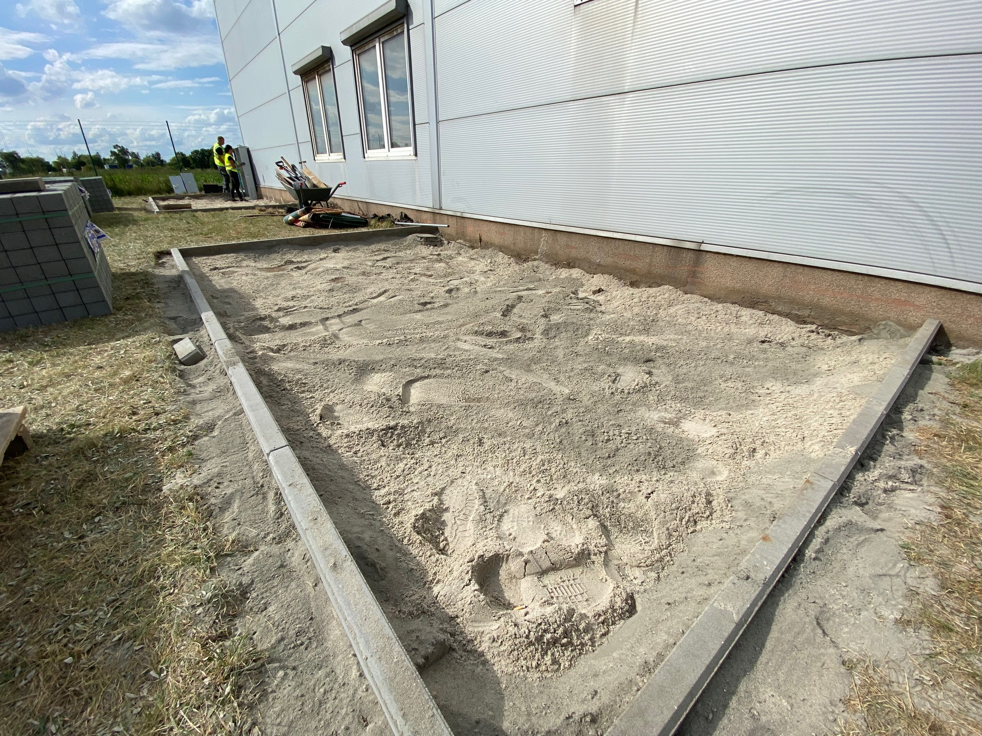 Przygotowanie podłoża pod kostkę brukową przy hali przemysłowej, widoczne obrzeża z kostki, piasek oraz ślady butów roboczych na piasku. W tle pracownicy i narzędzia.