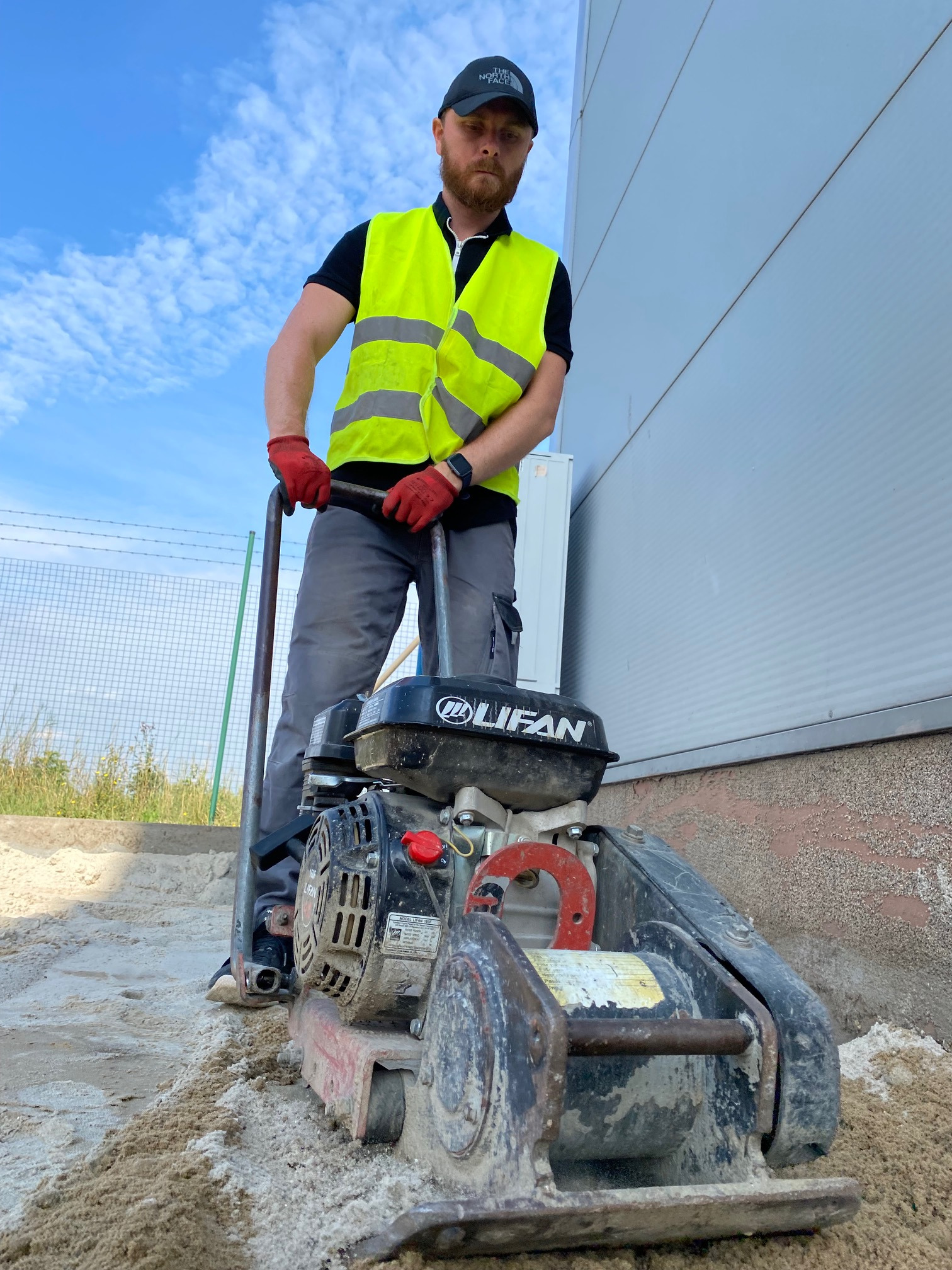Pracownik w kamizelce odblaskowej zagęszcza piasek zagęszczarką płytową Lifan przed budynkiem hali przemysłowej.