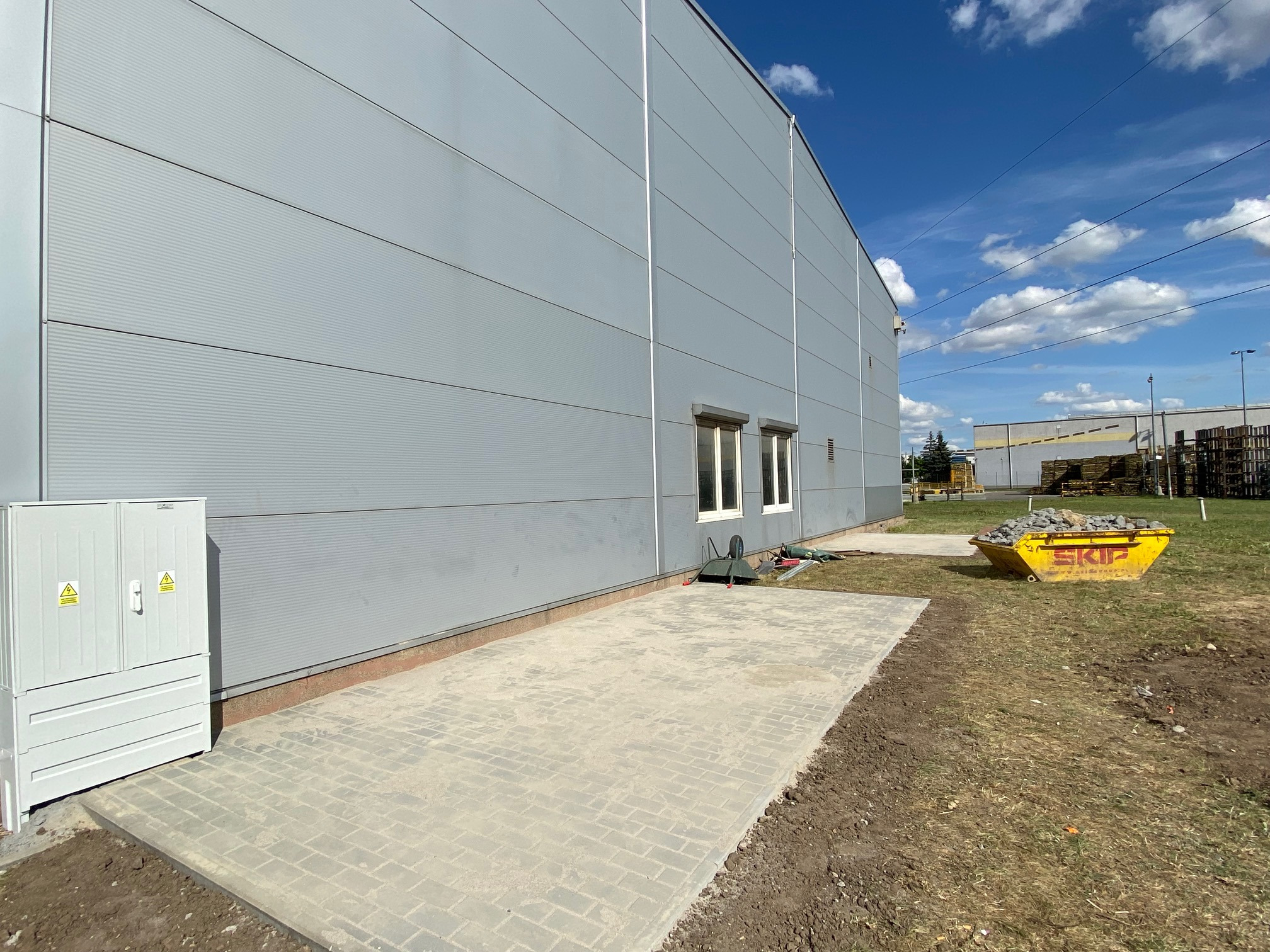 Wykonana nawierzchnia z kostki brukowej przy hali przemysłowej, widoczny żółty kontener na gruz, szafka elektryczna i fragment elewacji hali z oknami.
