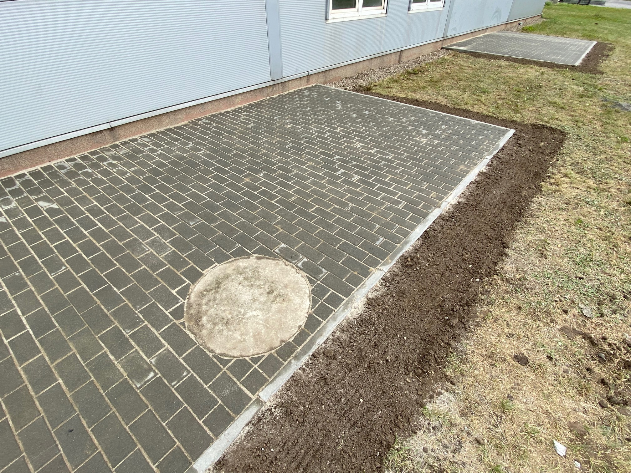 Ułożona kostka brukowa z okrągłą studzienką rewizyjną przy hali przemysłowej, widoczne obrzeża i przygotowane podłoże pod trawnik.