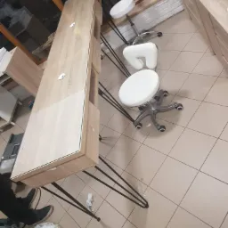 Długi, wąski blat z jasnego drewna z szklaną obwódką, wsparty na czarnych, metalowych nogach typu hairpin, w tle białe krzesła obrotowe i ceglana ściana.