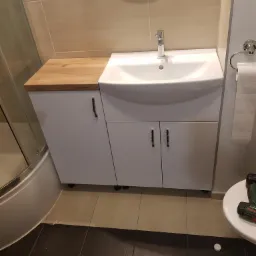 Biała szafka łazienkowa z umywalką i blatem z jasnego drewna, widoczny klucz nastawny i wiertarka na podłodze, odzwierciedlenie osoby w lustrze.