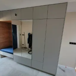 Szara zabudowa meblowa z lustrami i siedziskiem obok okna, widoczna klatka schodowa w tle.