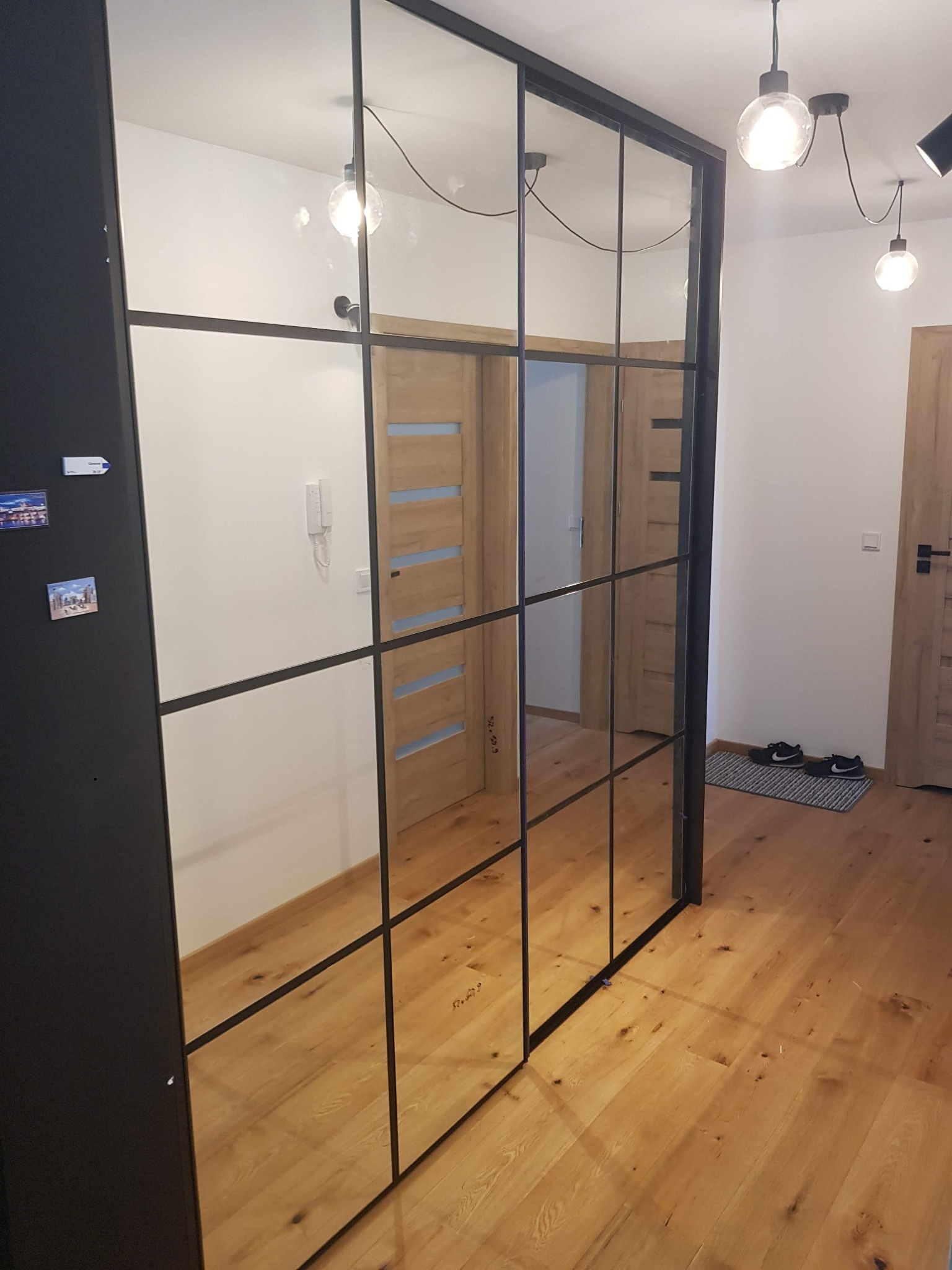 Duża szafa wnękowa z przesuwnymi drzwiami lustrzanymi z czarnymi szprosami, widoczne odbicie wnętrza mieszkania z drewnianymi drzwiami i podłogą.