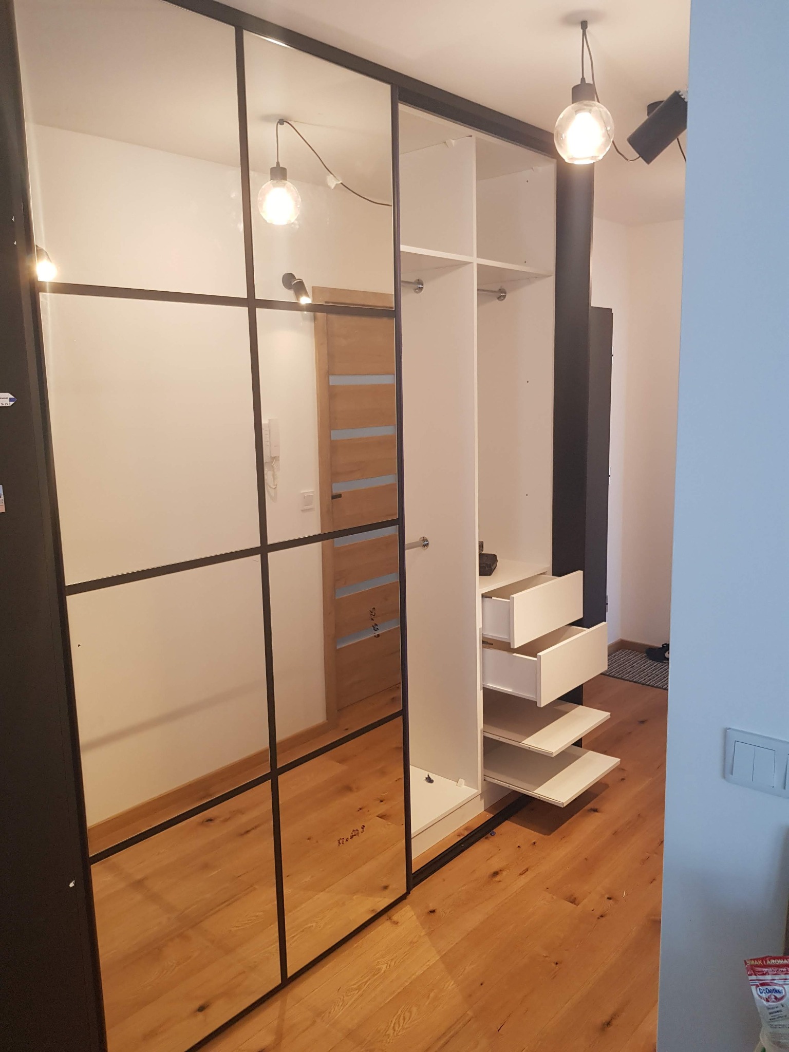 Nowoczesna szafa wnękowa z lustrzanymi drzwiami w czarnej ramie, widoczne otwarte półki i szuflady, oświetlenie punktowe i wiszące.