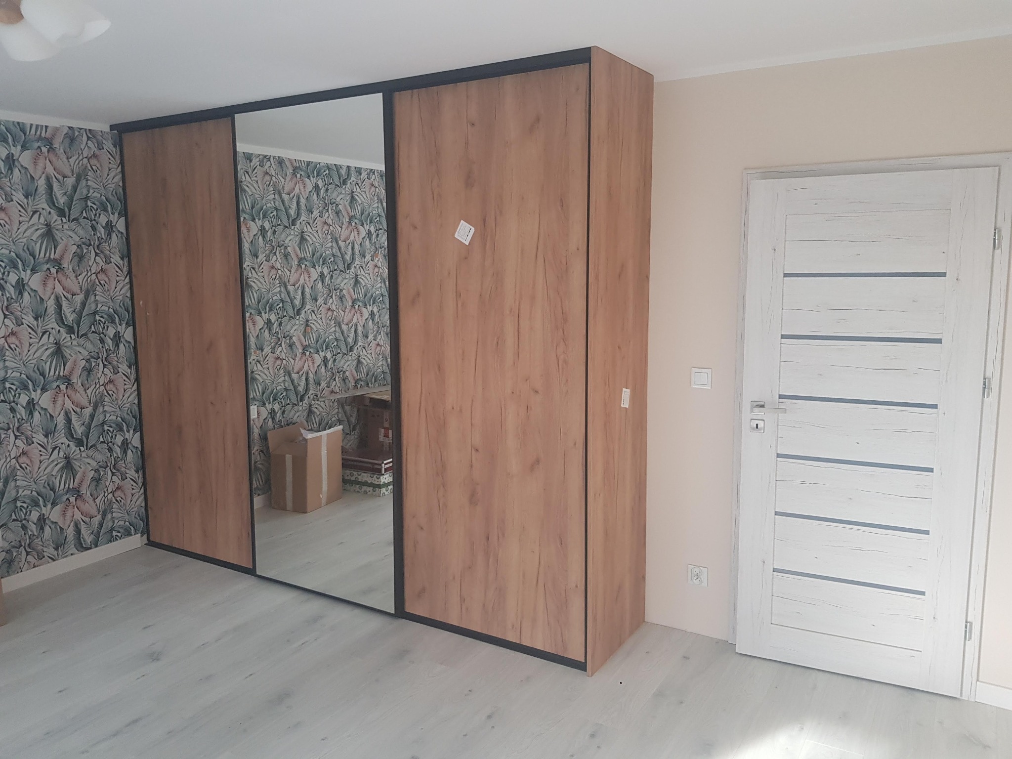 Trzydrzwiowa szafa przesuwna z lustrem i drewnopodobnymi frontami w pokoju z tapetą w roślinny wzór, widoczny karton w odbiciu lustrzanym.