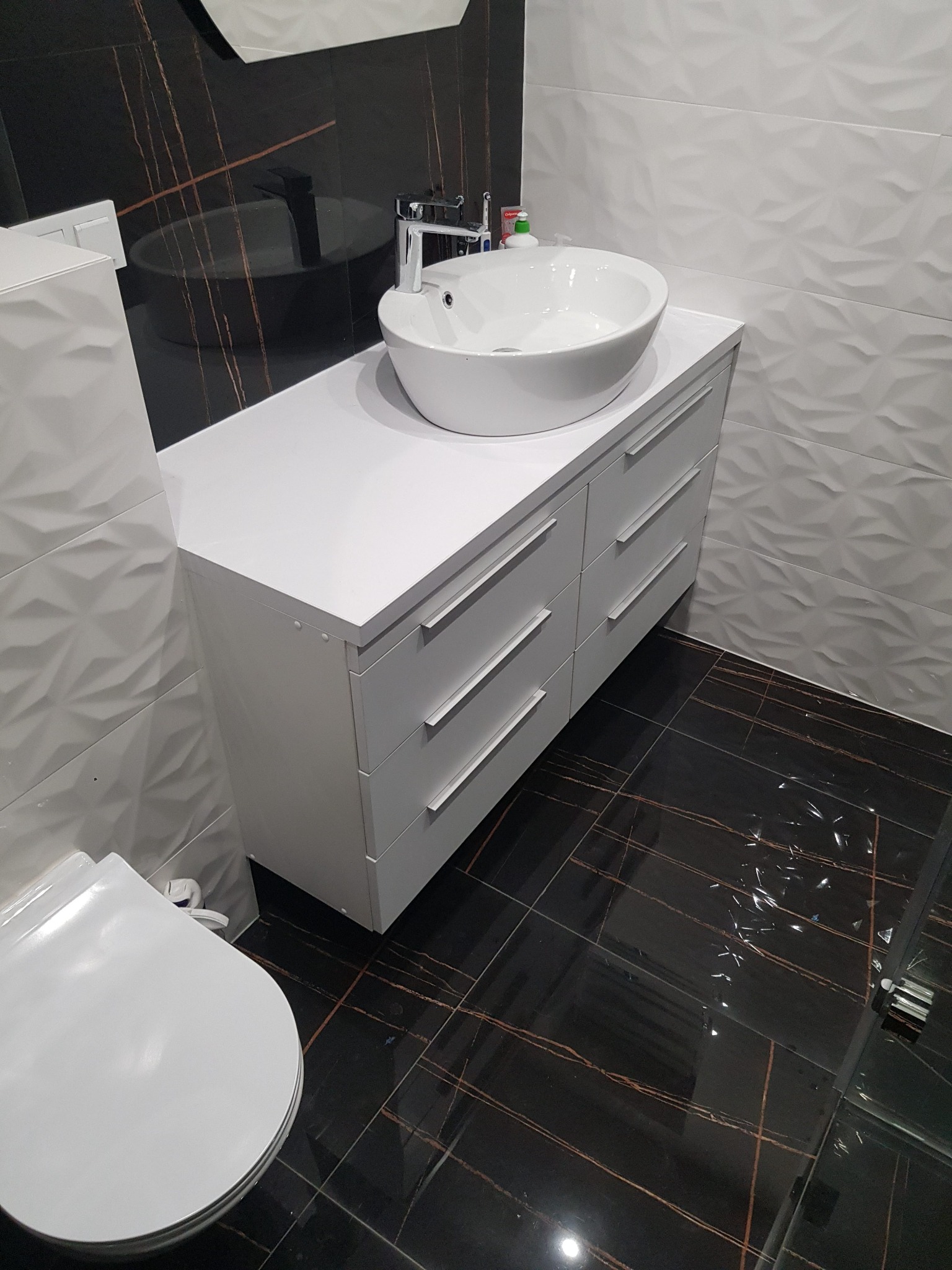 Nowoczesna łazienka z białą umywalką nablatową i szafką z szufladami, czarne płytki podłogowe z brązowymi żyłami, białe płytki ścienne z geometrycznym wzorem.