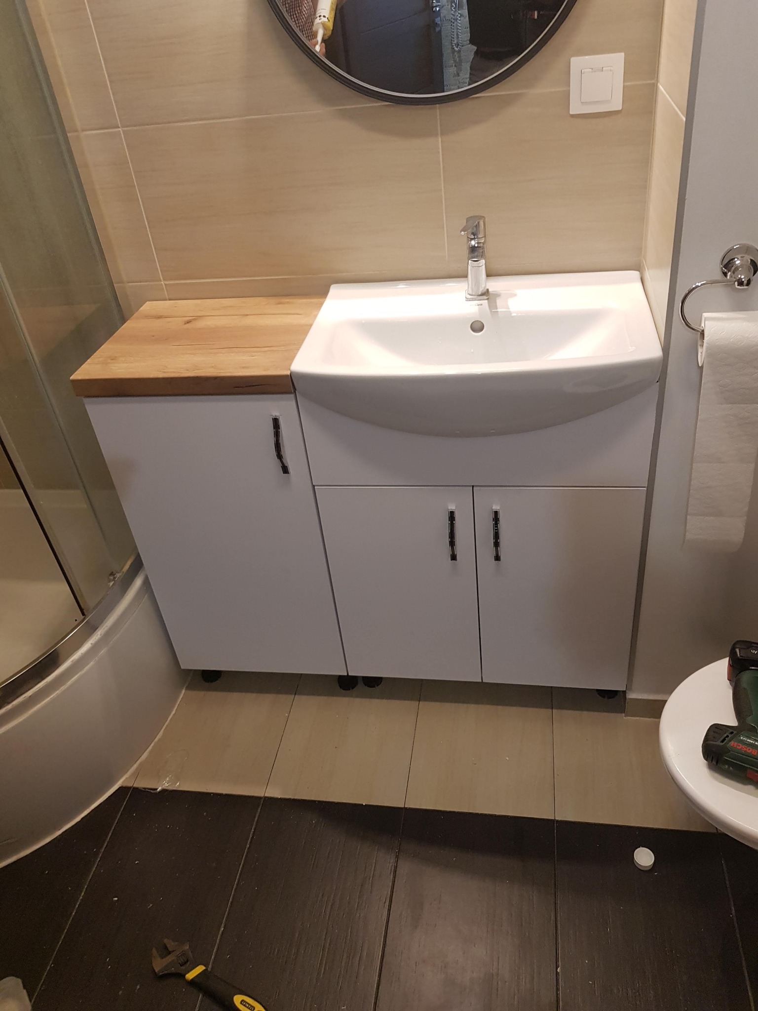 Biała szafka łazienkowa z umywalką i blatem z jasnego drewna, widoczny klucz nastawny i wiertarka na podłodze, odzwierciedlenie osoby w lustrze.