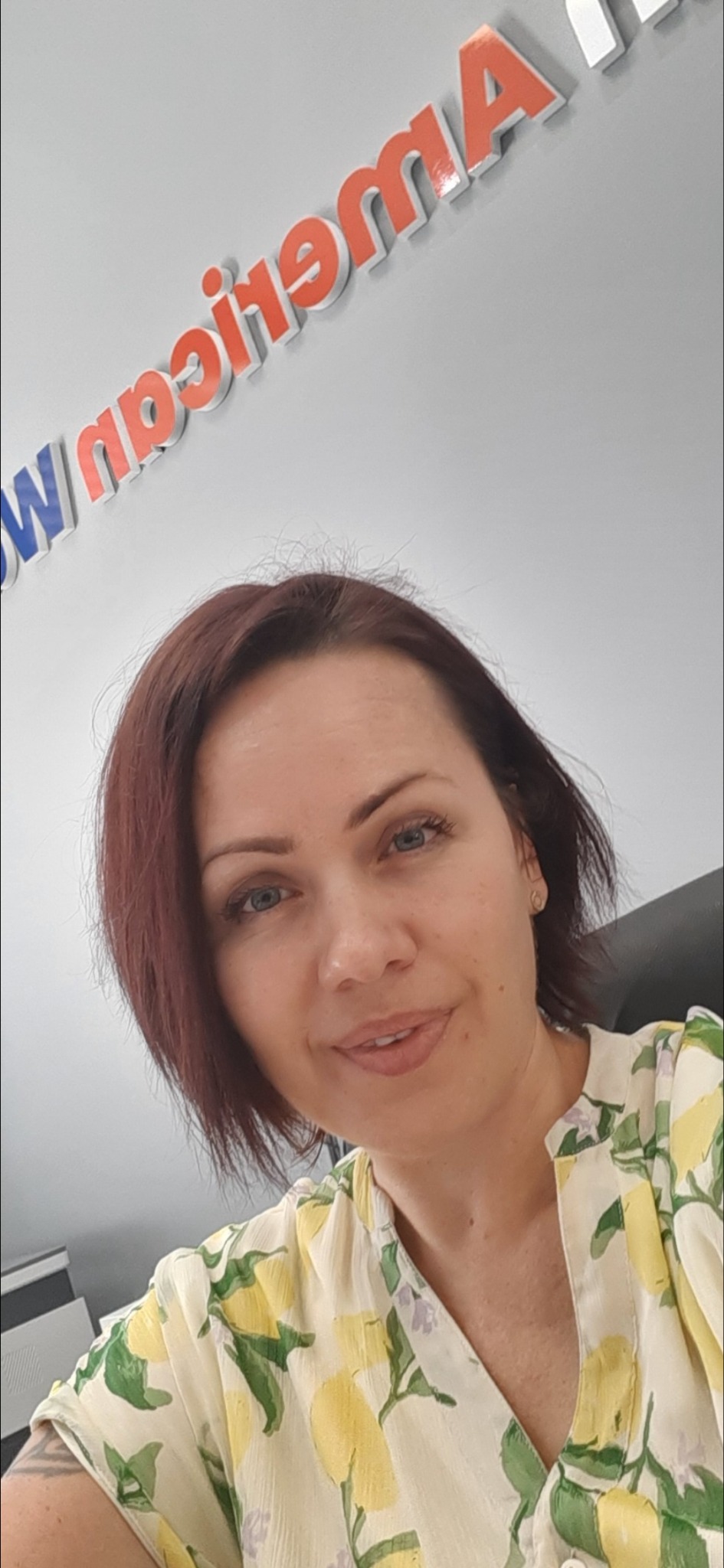 Portret kobiety z krótkimi, ciemnymi włosami, ubranej w bluzkę w cytrynowy wzór, na tle ściany z napisem 'American'.