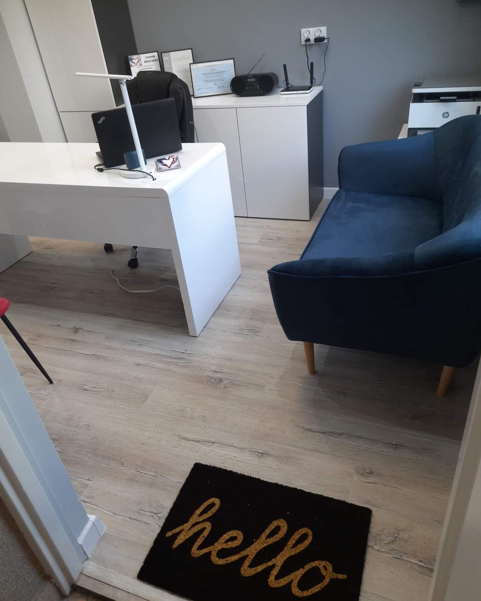 Nowoczesne biuro z białym biurkiem, laptopem, lampką, czarną marynarką na krześle, niebieską sofą i dywanikiem z napisem 'hello' na podłodze.
