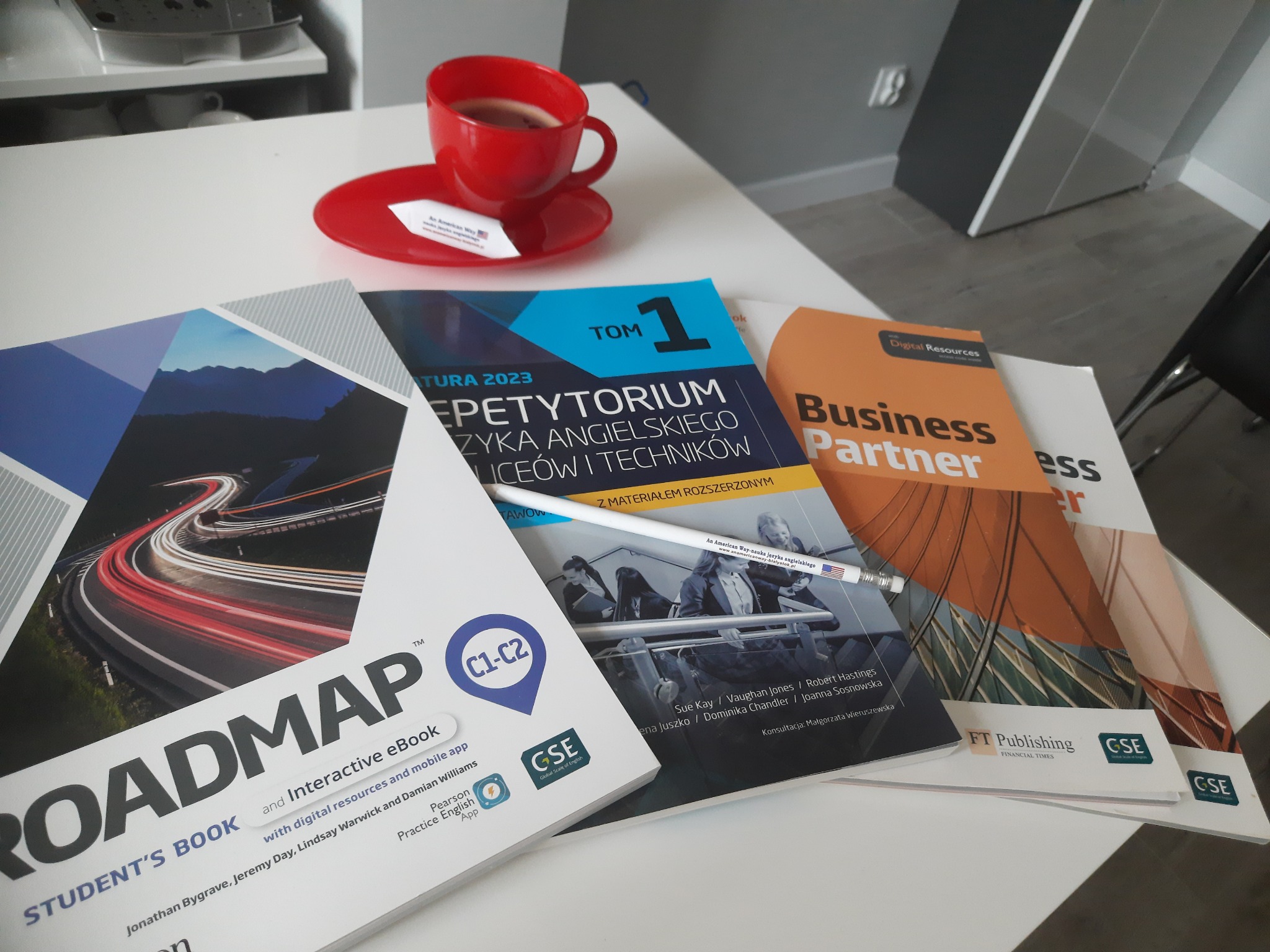 Stos podręczników do nauki języka angielskiego, w tym 'Roadmap', 'Repetytorium' i 'Business Partner', ułożonych na białym stole obok czerwonej filiżanki z kawą i długopisu z logo szkoły językowej.