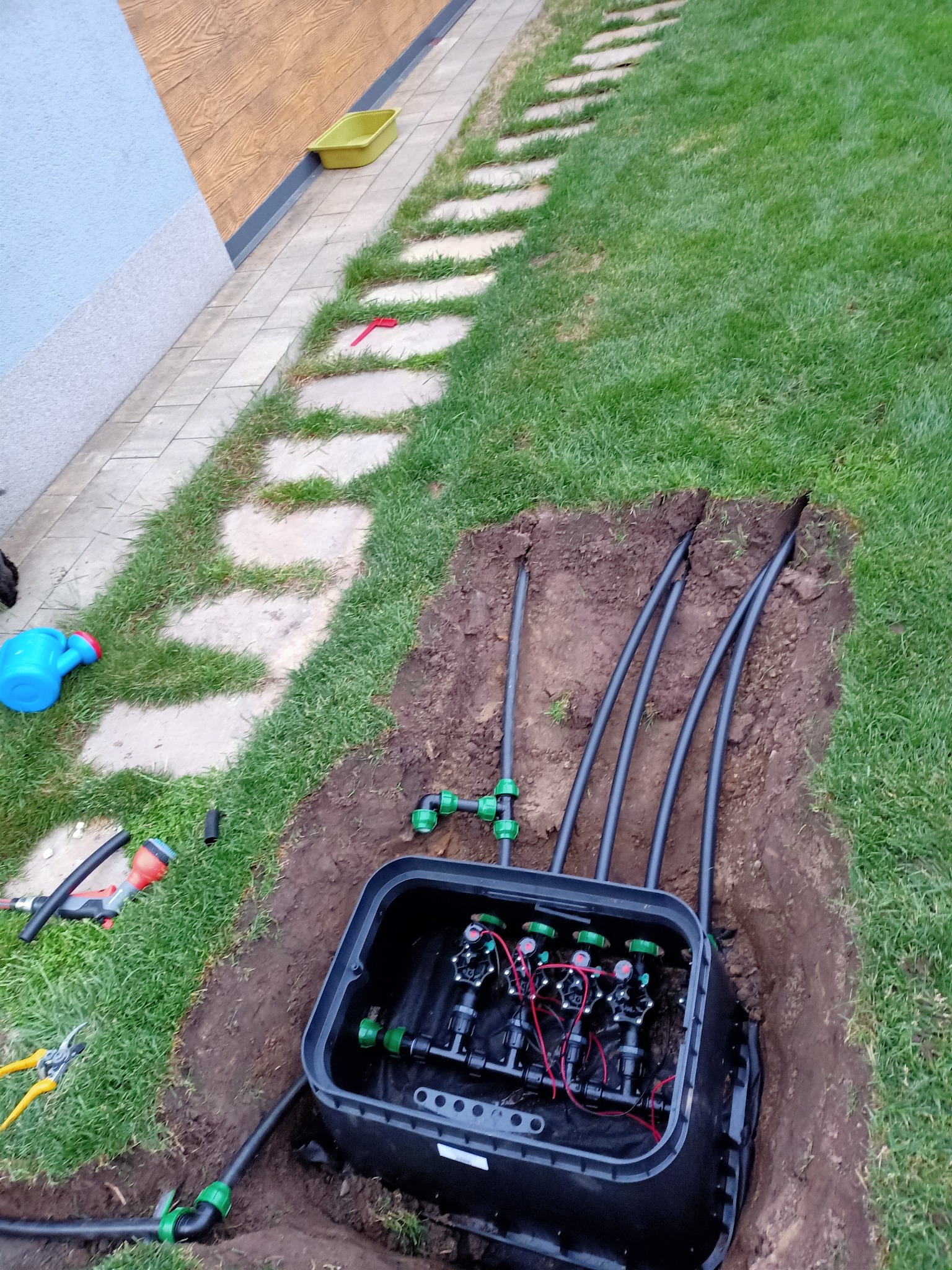 Instalacja automatycznego systemu nawadniania trawnika z podziemną skrzynką sterującą, widoczne rury doprowadzające wodę i złącza, obok narzędzia ogrodnicze i konewka.