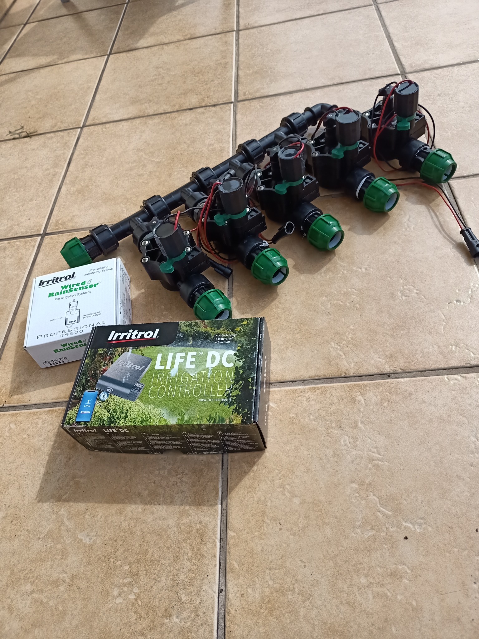Zestaw elektrozaworów do nawadniania z kontrolerem Irritrol LIFE DC i czujnikiem deszczu RS500, rozłożony na kafelkach podłogowych.