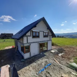 Surowy stan domu jednorodzinnego z ciemną dachówką i drewnianymi oknami, widoczne bloczki konstrukcyjne, teren wokół w trakcie przygotowania, w tle pagórkowaty krajobraz.