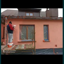 Pracownik na rusztowaniu maluje elewację domu natryskowo, okna zabezpieczone folią, widoczny kubeł z farbą. Ujęcie z dołu.