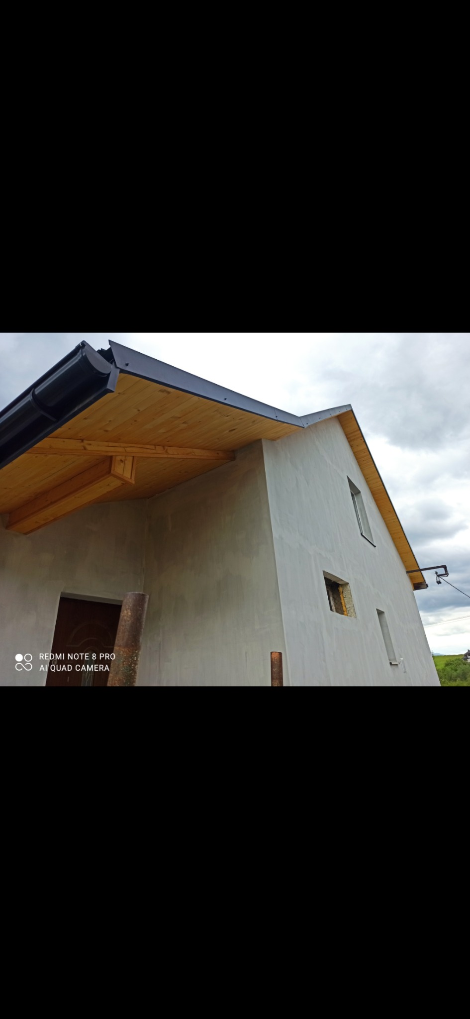 Surowy budynek jednorodzinny z szarą elewacją w trakcie wykańczania, widoczne deskowanie podbitki dachowej, fragment krajobrazu w tle.