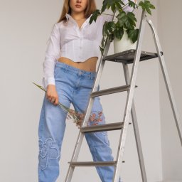 Michał Borowczyk - Młoda kobieta w białej koszuli i jeansach z aplikacjami, pozująca z drabinką i rośliną doniczkową na białym tle, trzymając w dłoni bukiecik suszonych kwiatów.