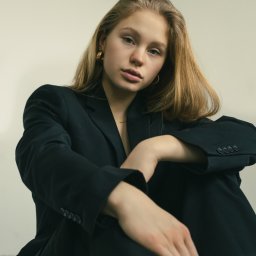 Michał Borowczyk - Portret młodej kobiety w czarnym, oversize'owym garniturze, z delikatną złotą biżuterią, siedzącej na podłodze i patrzącej w obiektyw z lekko pochyloną głową.