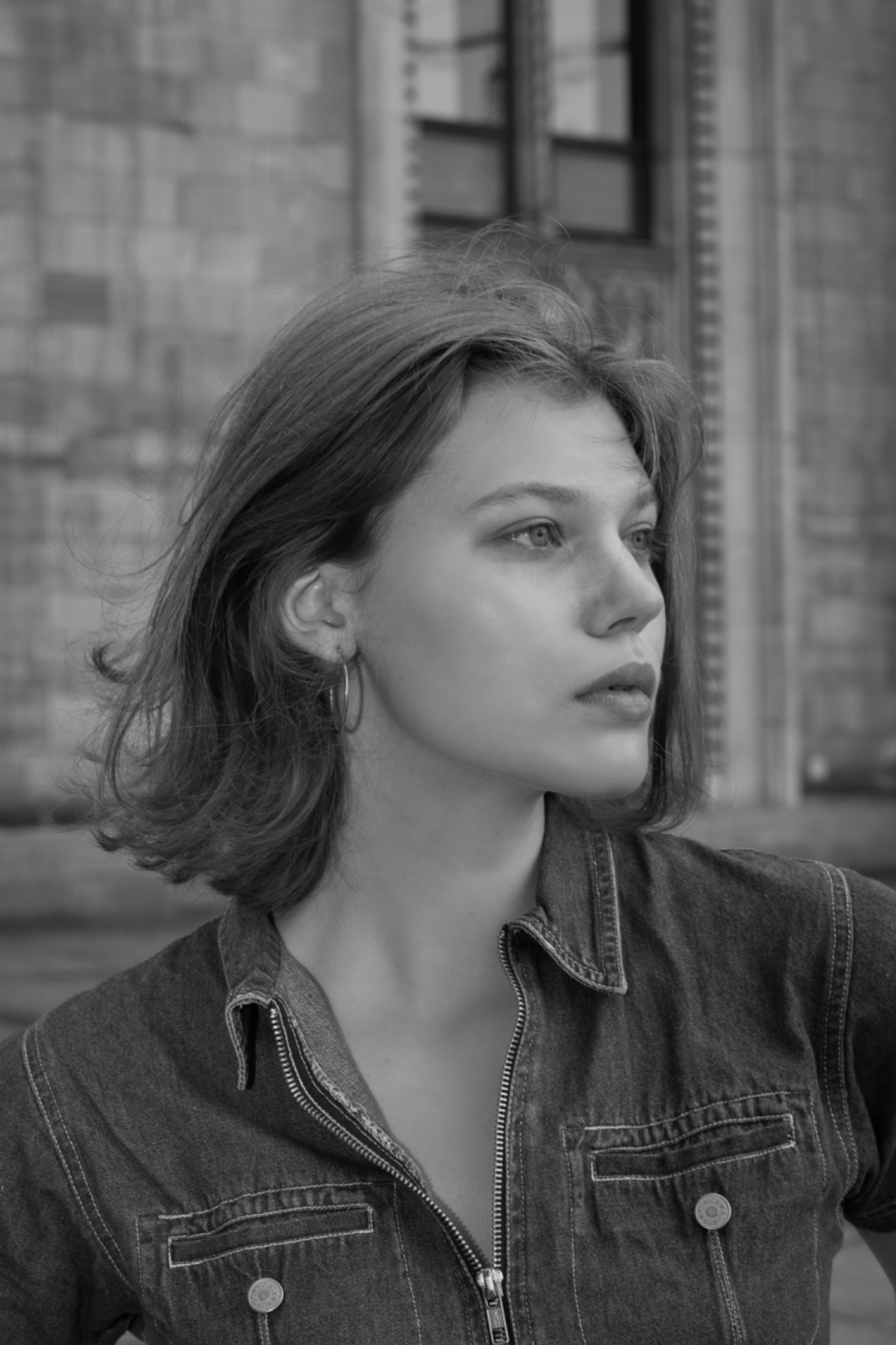 Czarno-białe portretowe ujęcie młodej kobiety z krótkimi włosami, ubranej w dżinsową kurtkę, z budynkiem w tle.