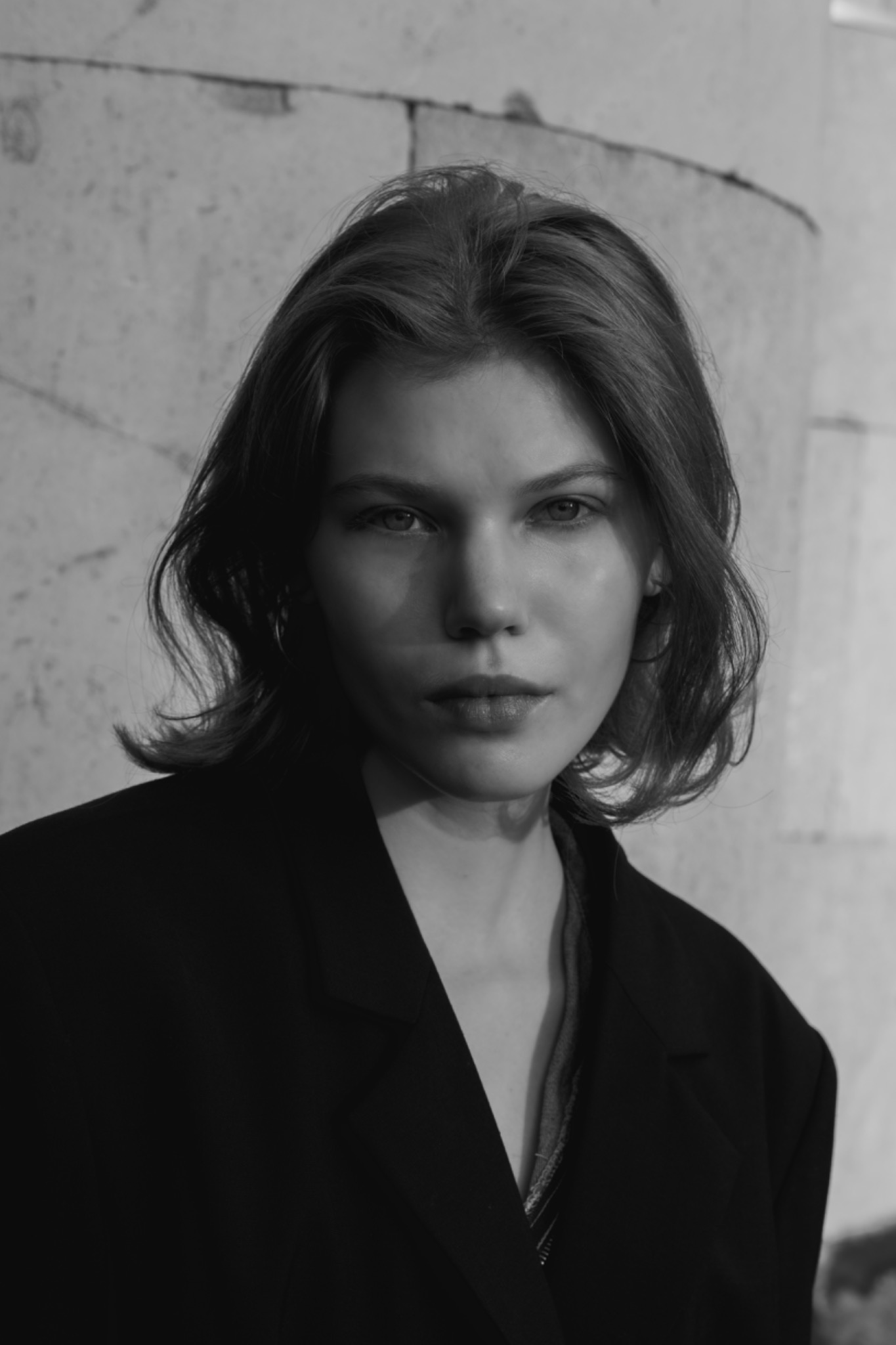 Portret młodej kobiety w czarno-białej tonacji, z delikatnym makijażem i rozpuszczonymi włosami, ubrana w czarną marynarkę na tle jasnej, teksturowanej ściany.
