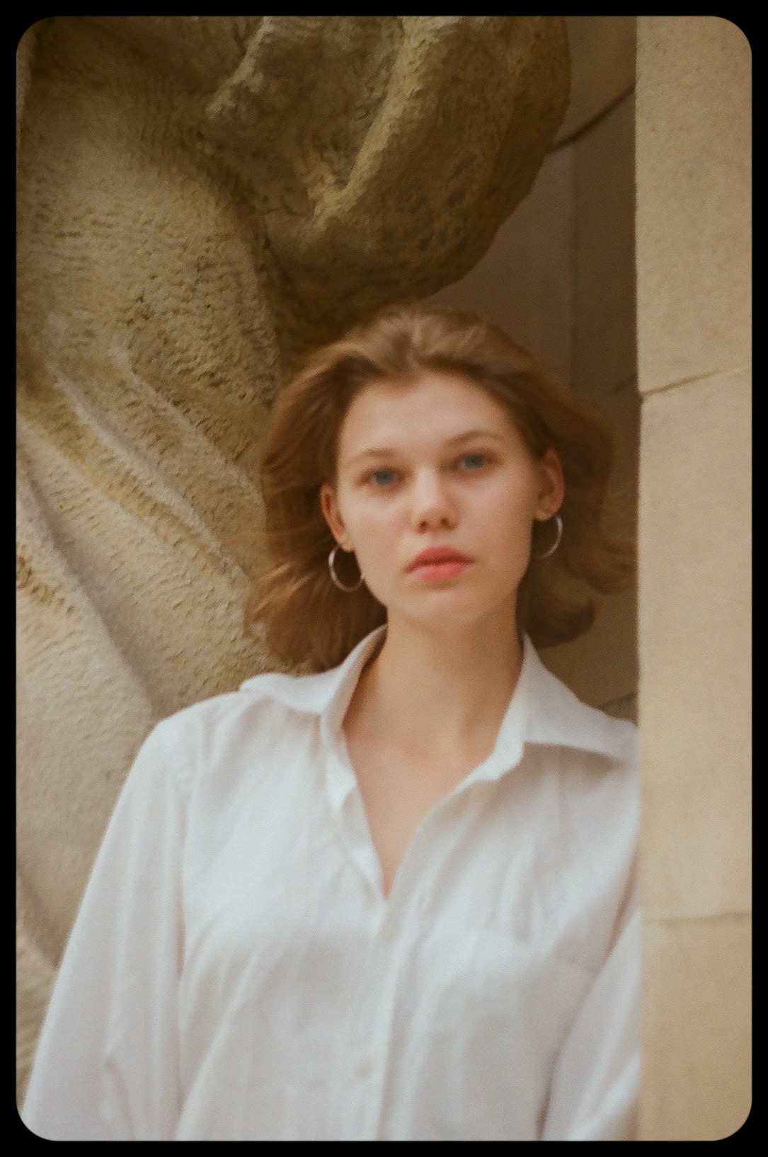 Portret młodej kobiety w białej koszuli na tle kamiennej rzeźby i kolumny, z kolczykami kołami i rozpuszczonymi włosami.