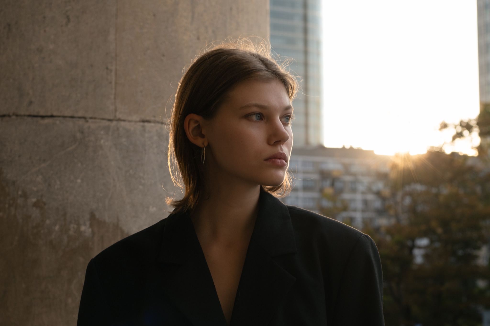 Portret młodej kobiety w czarnym żakiecie, z kolczykiem w uchu, oświetlonej zachodzącym słońcem na tle miejskiego krajobrazu.
