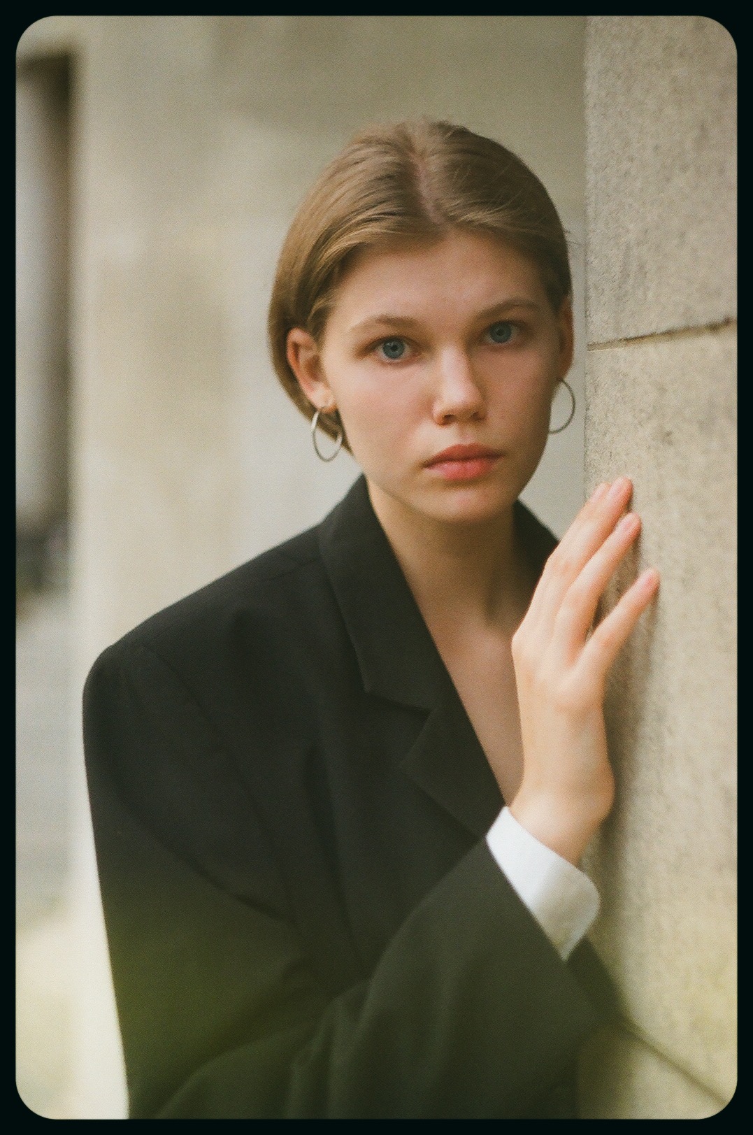 Portret młodej kobiety w czarnej marynarce, opierającej dłoń o kamienną ścianę, z delikatnym makijażem i srebrnymi kolczykami kołami.