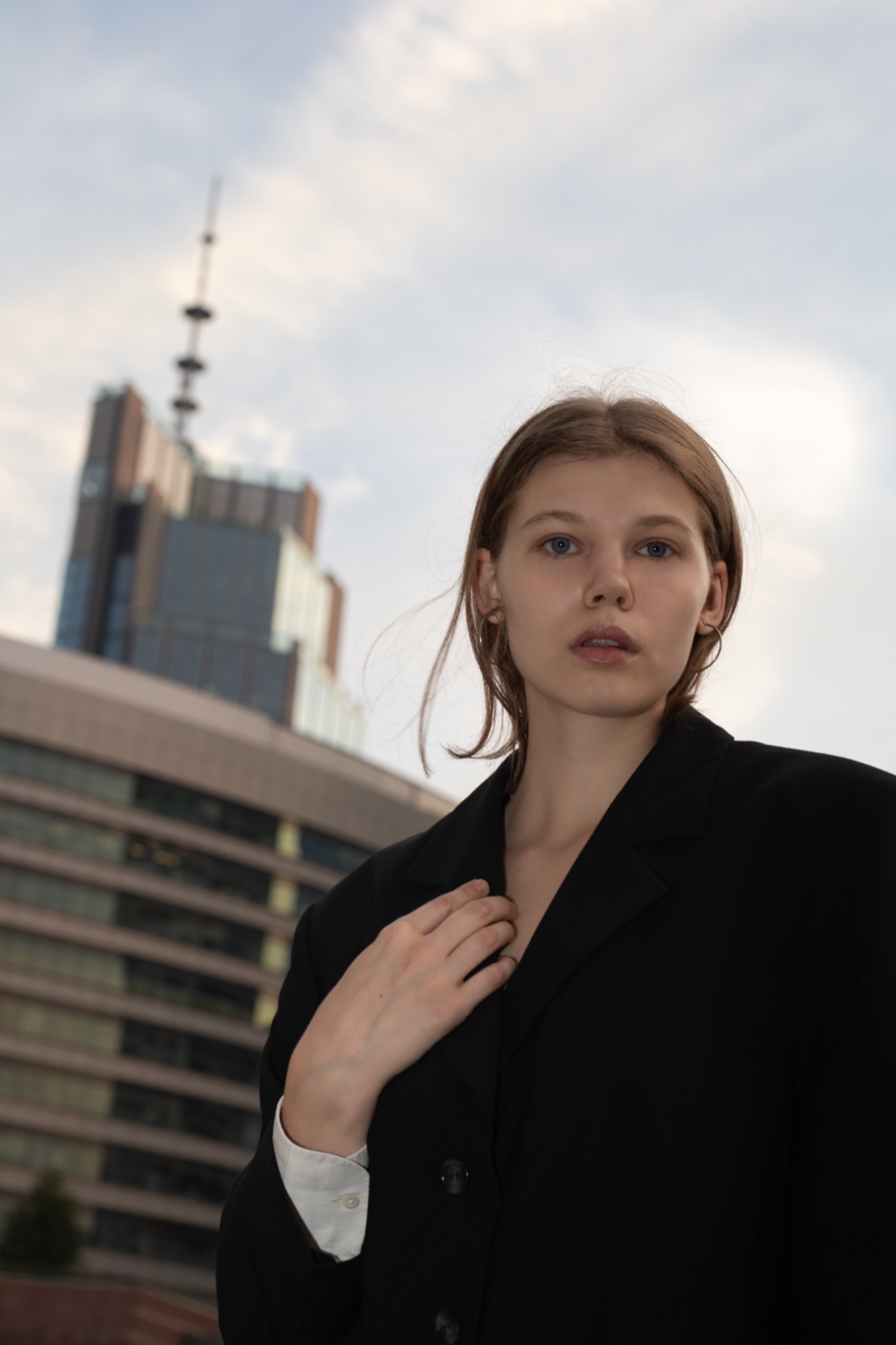 Portret młodej kobiety w czarnej marynarce na tle miejskiego krajobrazu z wieżowcem i pochmurnym niebem.