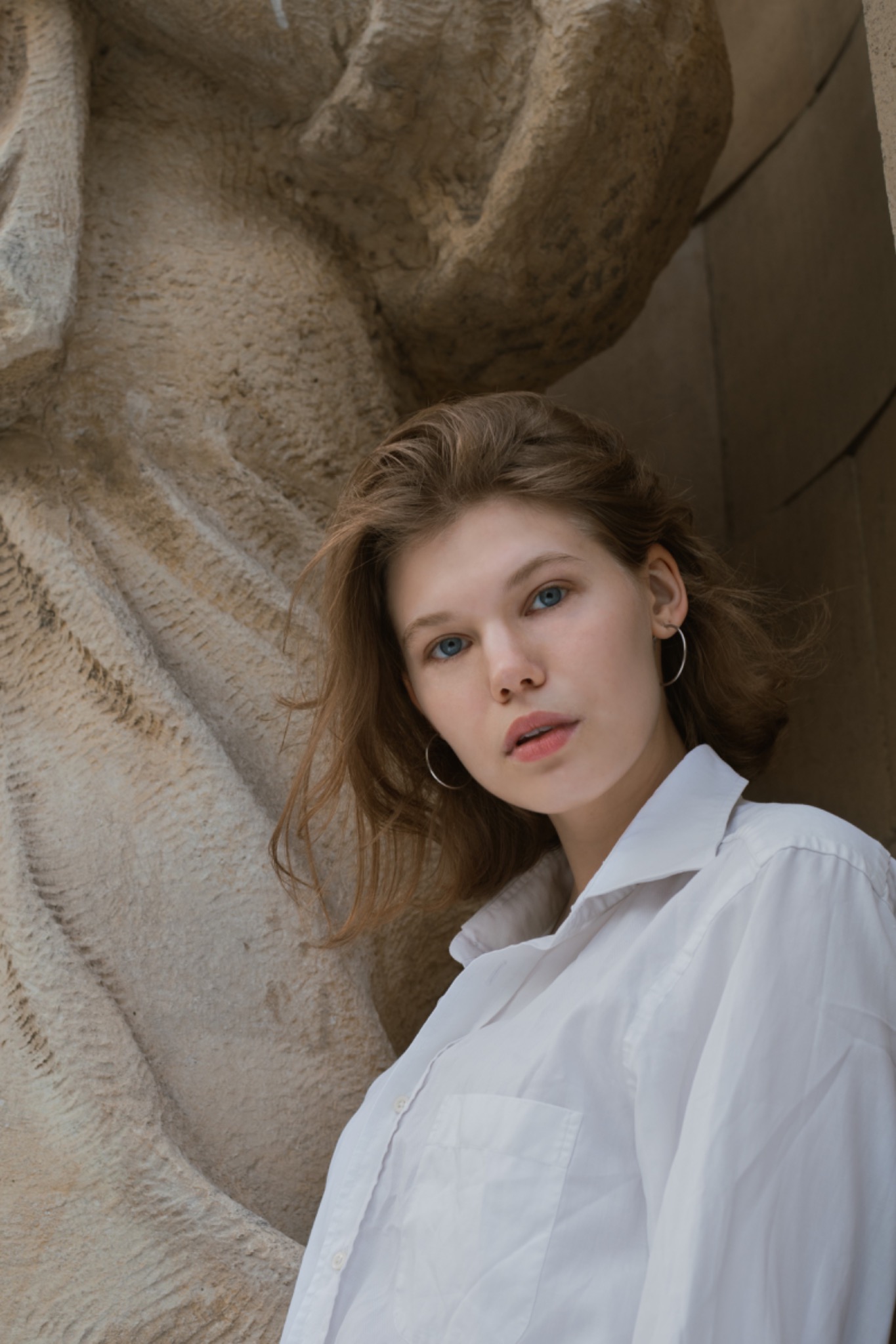 Portret młodej kobiety w białej koszuli na tle kamiennej rzeźby, z delikatnym makijażem i srebrnymi kolczykami-kołami.