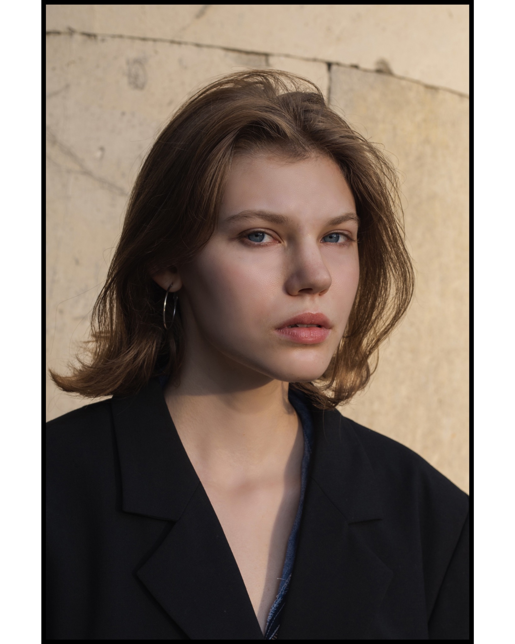 Portret młodej kobiety z krótkimi brązowymi włosami i niebieskimi oczami, ubrana w czarną marynarkę, pozująca na tle jasnej, kamiennej ściany.