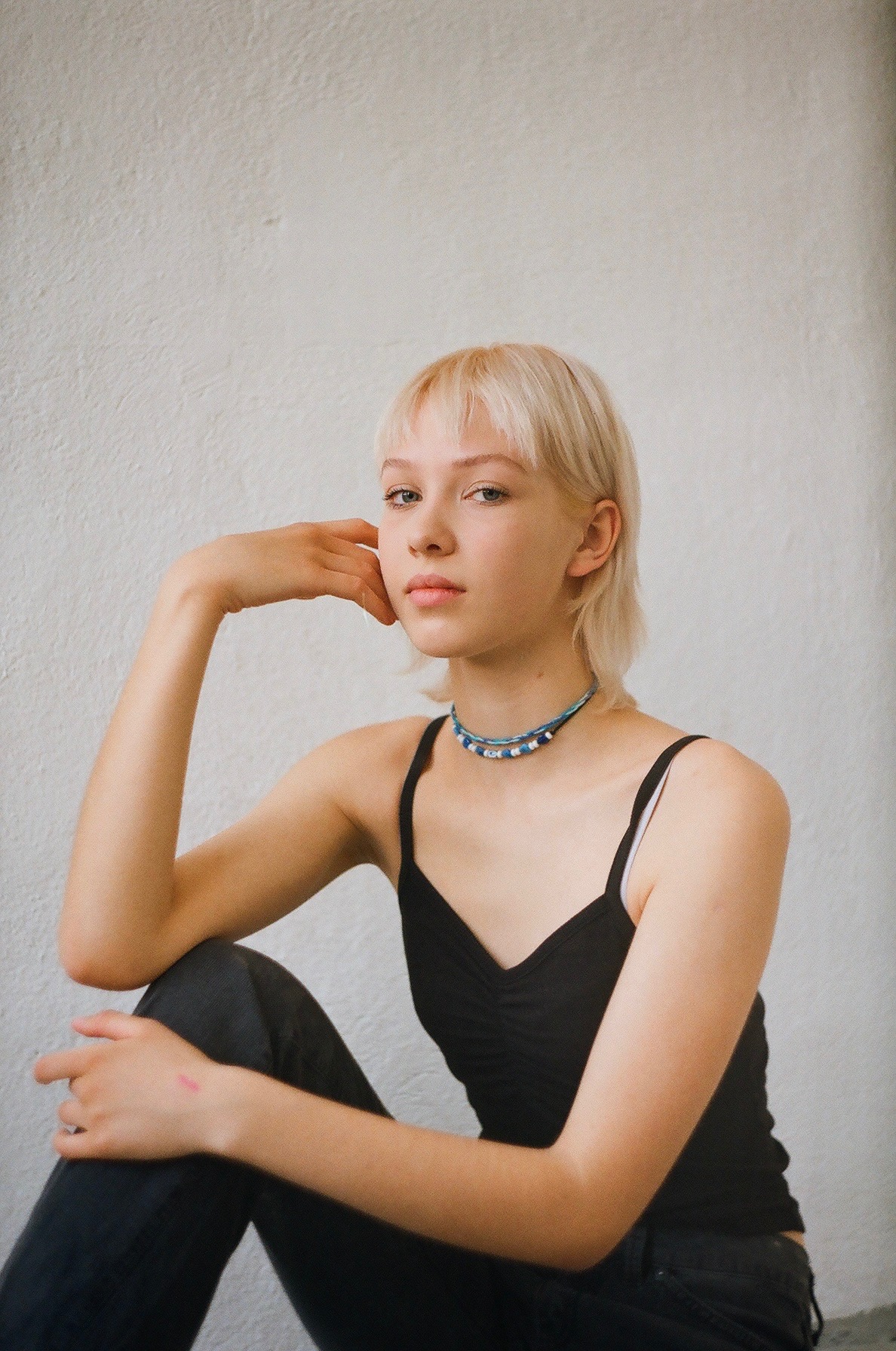 Portret młodej kobiety z krótkimi blond włosami, siedzącej na podłodze i opierającej głowę na dłoni, ubrana w czarny top na ramiączkach i dżinsy, z delikatnym makijażem i naszyjnikiem z koralików...