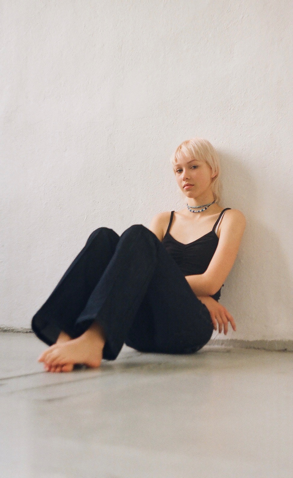 Młoda kobieta z blond włosami siedzi boso na podłodze oparta o białą ścianę, ubrana w czarny top na ramiączkach i ciemne spodnie, z delikatnym naszyjnikiem.