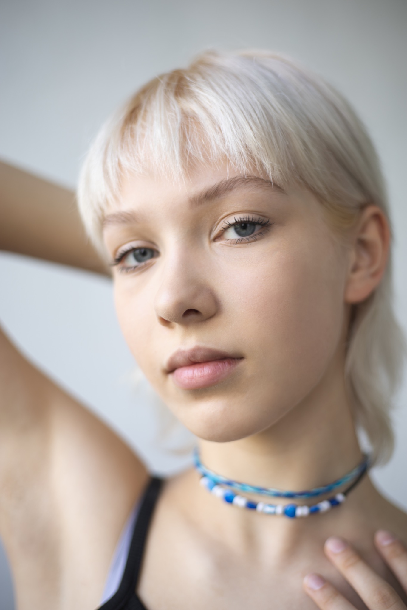 Portret młodej kobiety z platynowymi włosami, błękitnymi oczami i naszyjnikiem z koralików, pozującej na jasnym tle.