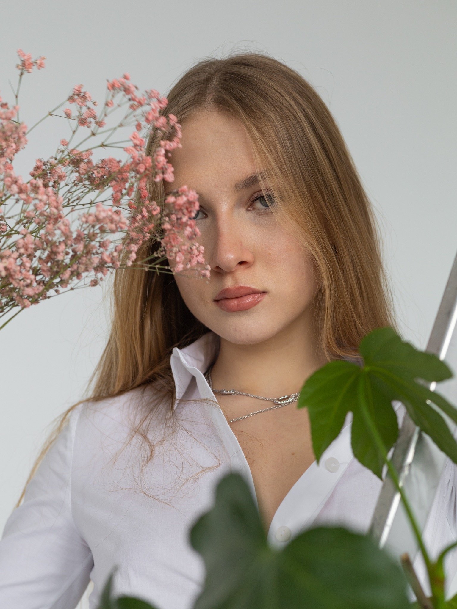 Portret młodej kobiety w białej koszuli, z delikatnymi różowymi kwiatami zasłaniającymi część twarzy i zielonym liściem w prawym dolnym rogu, na jasnym tle.