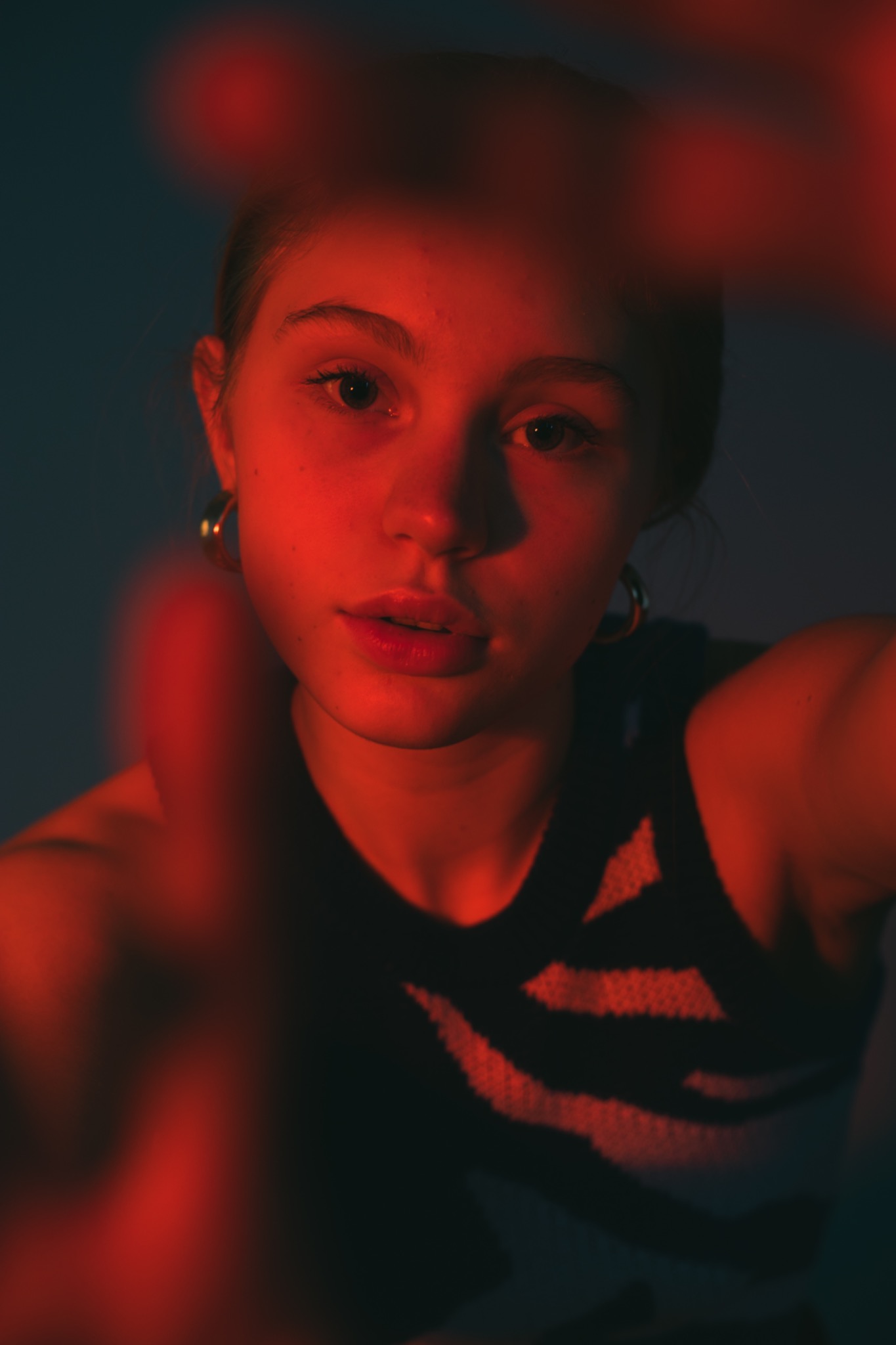 Portret młodej kobiety z kolczykami, oświetlonej czerwonym światłem, z rozmazanymi dłońmi na pierwszym planie, tworzącymi artystyczny efekt głębi.