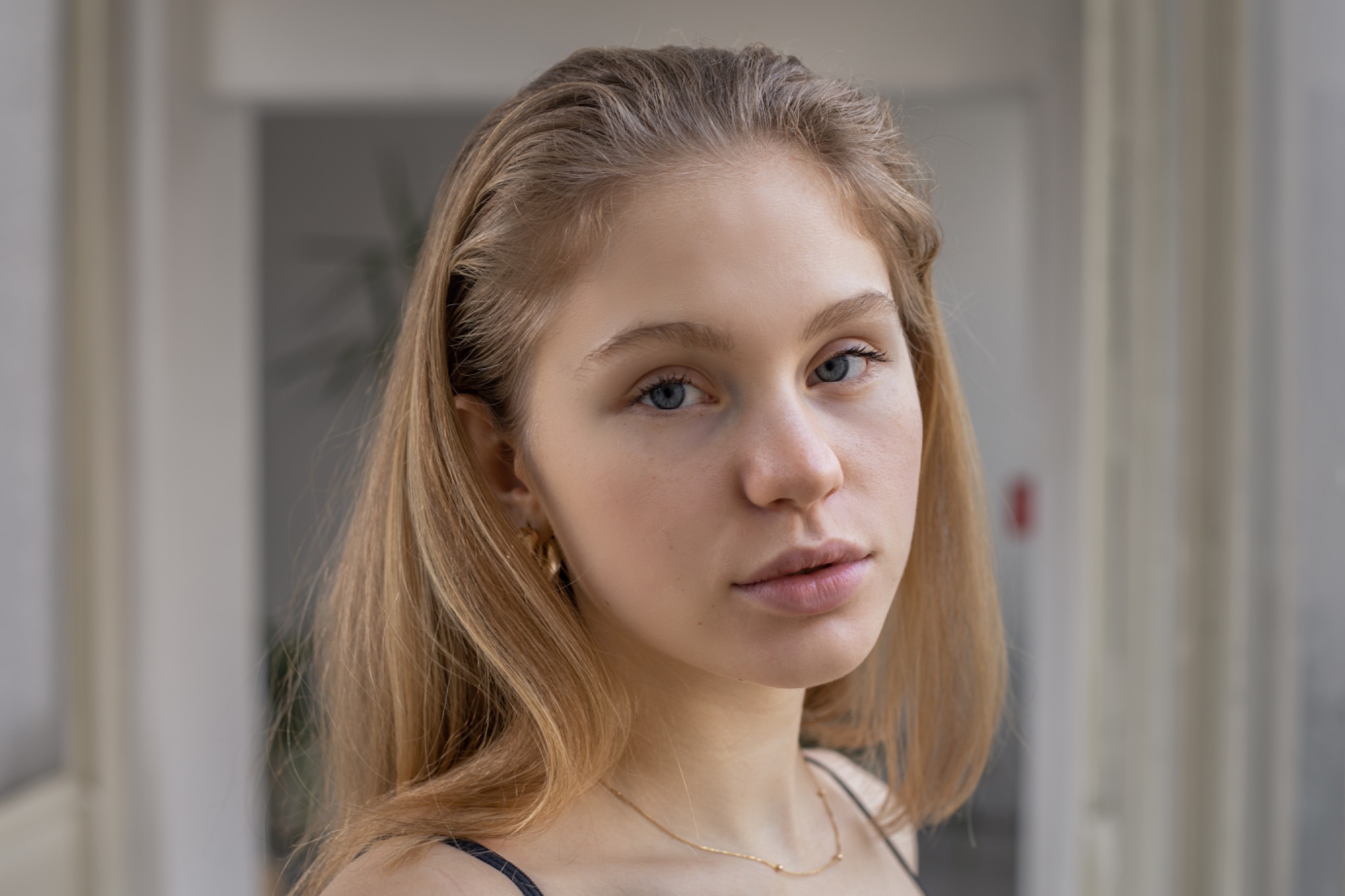 Portret młodej kobiety o jasnych włosach i niebieskich oczach, z delikatną złotą biżuterią, na rozmytym neutralnym tle.