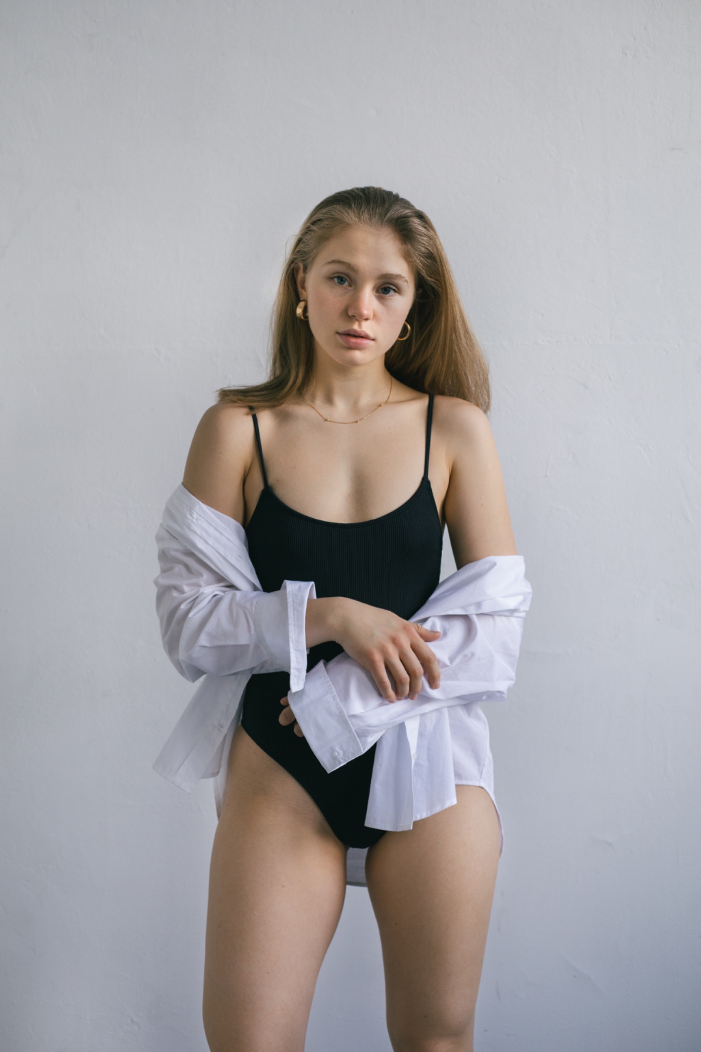 Portret kobiety w czarnym body i rozpiętej białej koszuli na tle jasnej ściany, z delikatną złotą biżuterią.