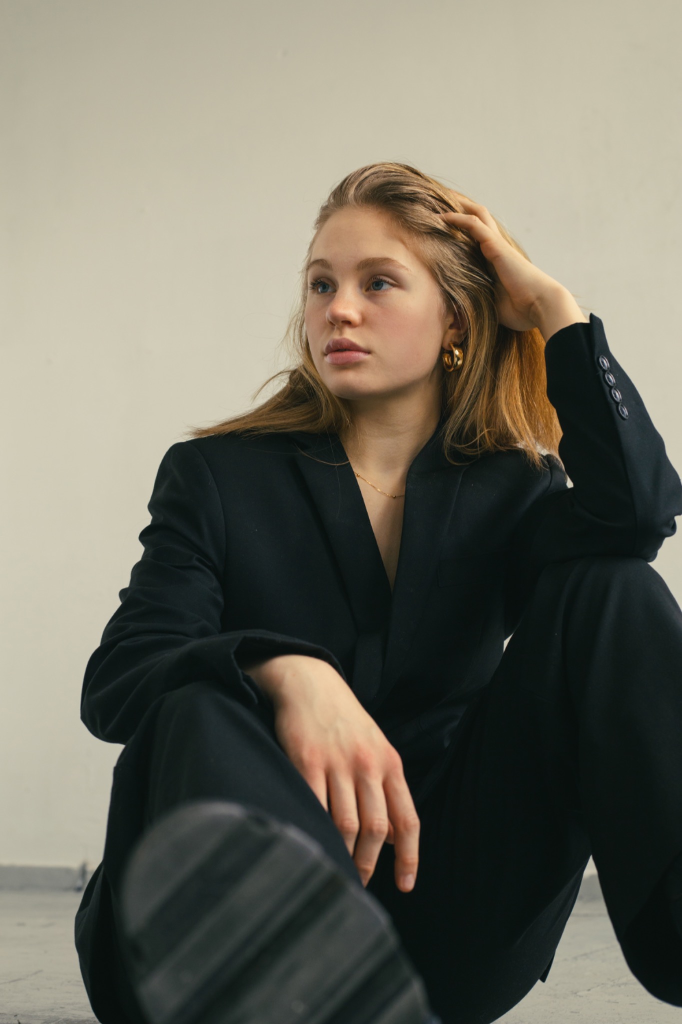 Młoda kobieta w czarnym garniturze siedzi na podłodze, zamyślona, z dłonią we włosach i złotymi kolczykami kołami.