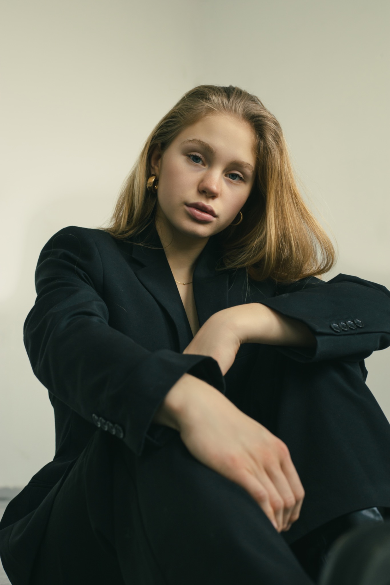 Portret młodej kobiety w czarnym, oversize'owym garniturze, z delikatną złotą biżuterią, siedzącej na podłodze i patrzącej w obiektyw z lekko pochyloną głową.