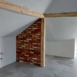 Wykończone poddasze w Chorzowie z fragmentem ściany wyłożonym dekoracyjną cegłą, drewnianymi belkami stropowymi i szarą wylewką podłogową.