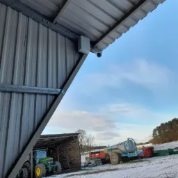 Zewnętrzna kamera monitoringu zamontowana na metalowej konstrukcji w pobliżu zabudowań gospodarczych i sprzętu rolniczego w zimowym krajobrazie.