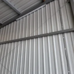 Wewnętrzna ściana hali z blachy trapezowej, widoczne kable elektryczne w peszlach, metalowa konstrukcja nośna i lampa liniowa LED.