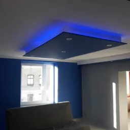 Nowoczesny pokój z podwieszanym, podświetlanym na niebiesko sufitem z wbudowanymi punktami świetlnymi, niebieskimi ścianami i widokiem na budynek z okna